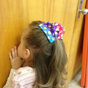 May include: A young girl with long brown hair wearing a pink and white striped shirt. Her hair is pulled back with a colorful polka dot hair tie with gold accents.