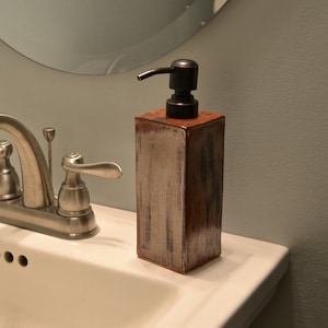 Puede incluir: Un lavabo de baño de porcelana blanca con un grifo plateado y dos dispensadores de jabón. Uno de los dispensadores es una caja de madera marrón con una bomba plateada, y el otro es un recipiente de metal plateado.