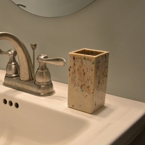 May include: A white porcelain bathroom sink with a silver faucet, a white soap dispenser with a silver pump, and a white toothbrush holder with brown and green flecks.