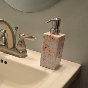 Op de afbeelding: Een witte keramische zeepdispenser met een kleurrijk gespikkeld patroon en een zilveren pomp. De dispenser staat op een wit badkamer wastafelblad naast een zilveren kraan.