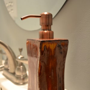 May include: A brown ceramic soap dispenser with a copper-coloured pump. The dispenser has a rectangular shape with curved sides and a glossy finish. The pump is angled and has a smooth, metallic appearance. The dispenser is on a white surface.