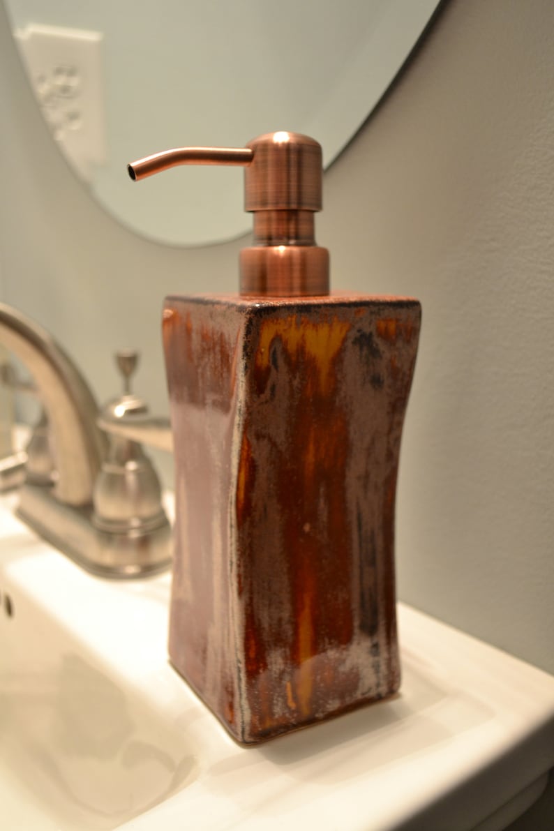 May include: A brown ceramic soap dispenser with a copper-colored pump. The dispenser has a rectangular shape with curved sides and a glossy finish. The pump is angled and has a smooth, metallic appearance. The dispenser is on a white surface.