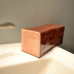 May include: A rectangular, handmade soap dish with a curved bottom. The dish is primarily brown with a lighter, pinkish-beige side. The name "A. Rollins" is inscribed on the side. The dish is sitting on a white surface.