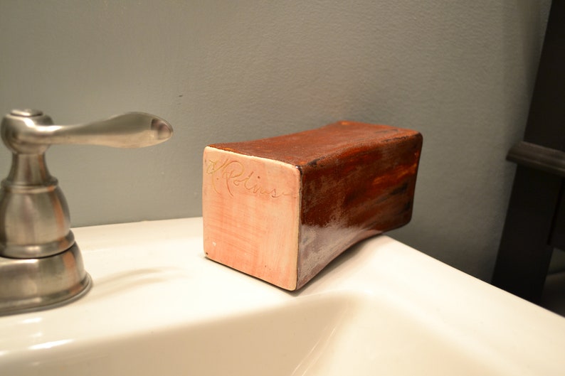 May include: A rectangular, handmade soap dish with a curved bottom. The dish is primarily brown with a lighter, pinkish-beige side. The name "A. Rollins" is inscribed on the side. The dish is sitting on a white surface.