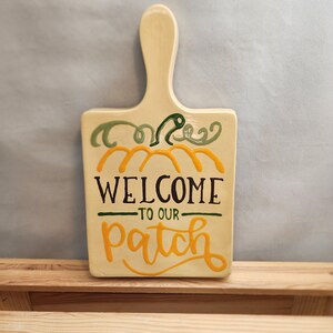 May include: A ceramic pumpkin-shaped sign with a handle. The sign is painted yellow with green vines and the words "Welcome to our patch" in black.
