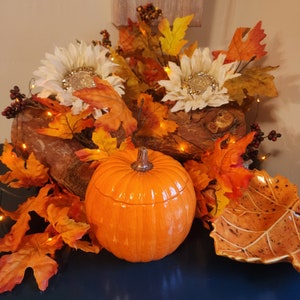 Peut inclure: Une citrouille en céramique avec un couvercle, décorée de feuilles d'automne et de lumières scintillantes. La citrouille est orange et a une tige sur le dessus. Les feuilles sont rouges, orange et jaunes. Les lumières scintillantes sont blanches et sont enfilées autour de la citrouille et des feuilles.
