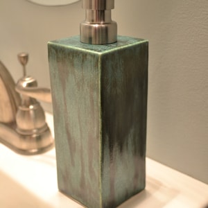 May include: A green ceramic soap dispenser with a silver pump. The dispenser is square shaped and has a textured surface.