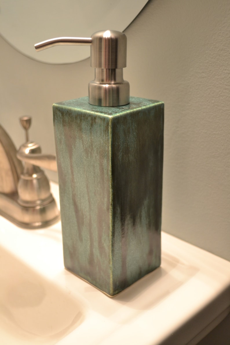 May include: A green ceramic soap dispenser with a silver pump. The dispenser is square shaped and has a textured surface.