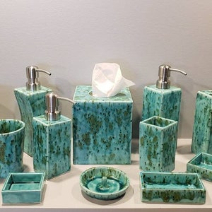 May include: A bathroom set of ceramic accessories in a teal green color with a speckled pattern. The set includes a soap dispenser, toothbrush holder, tumbler, tissue box cover, and various trays.