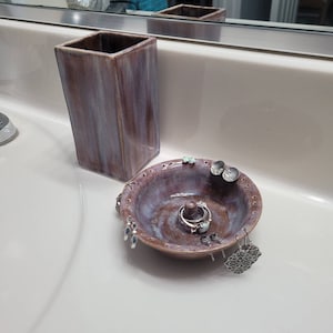 May include: A ceramic ring and earring dish with a purple and white glaze. The dish has a small center post for rings and holes around the edge for earrings. The dish is sitting on a white countertop next to a tall, rectangular ceramic container.