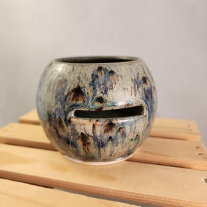 May include: A round ceramic planter with a slot opening. The planter has a speckled glaze in shades of blue, brown, and cream. The interior is dark. The planter sits on a white base.
