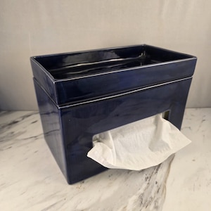 Puede incluir: Una caja rectangular para pañuelos de cerámica azul marino profundo con acabado brillante. La caja presenta una abertura rectangular ancha y centrada en la parte frontal, a través de la cual se ve un pañuelo de papel blanco asomando.