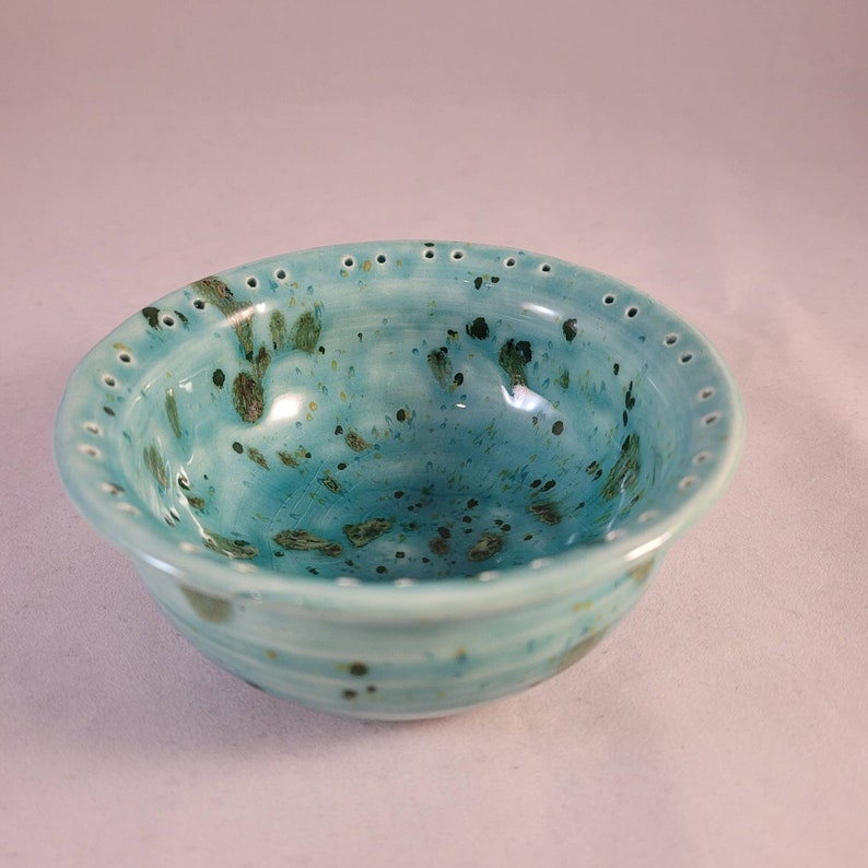 May include: A small, teal blue ceramic bowl with a speckled glaze and a row of holes around the rim.