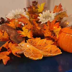 May include: A ceramic leaf-shaped dish with an orange and brown glaze, surrounded by autumn foliage, including maple leaves, and a small orange pumpkin.