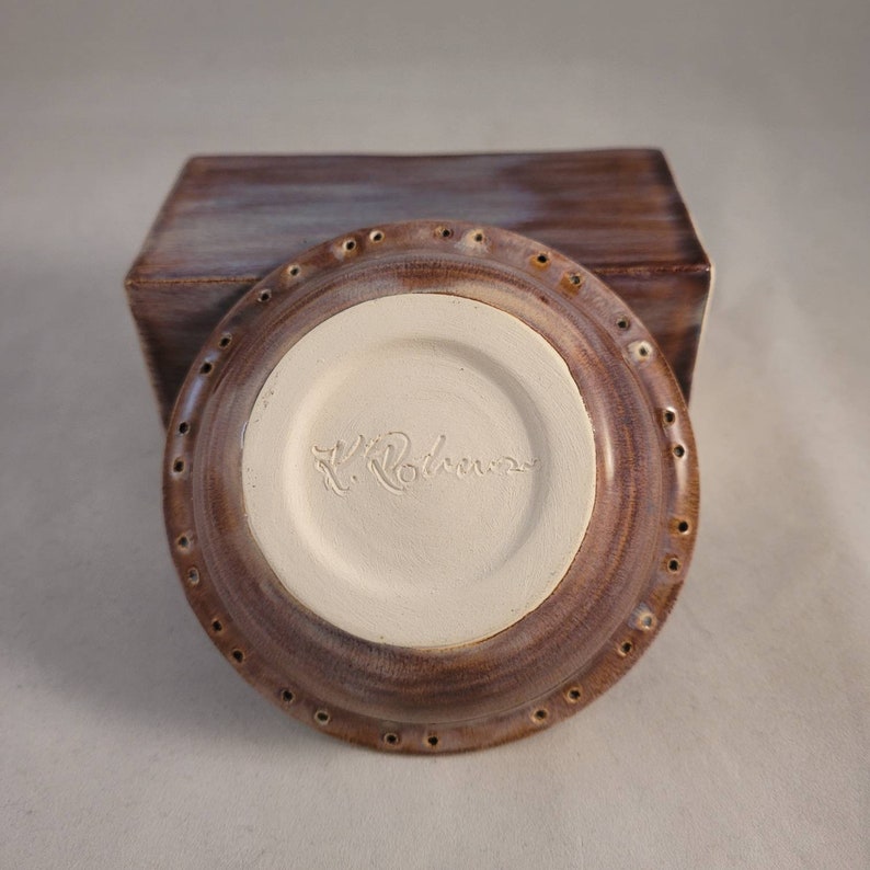 May include: A brown ceramic dish with a white interior and a ring of holes around the edge. The bottom of the dish is signed "L. Rolner".