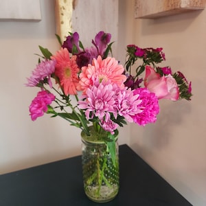 May include: A vibrant bouquet of pink, fuchsia, and peach-colored flowers in a clear glass jar. The arrangement includes gerbera daisies, chrysanthemums, and roses, creating a colorful floral display. The jar has a textured pattern.