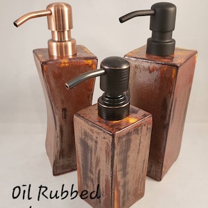 May include: Three decorative soap dispensers in copper, black, and oil-rubbed bronze finishes. The dispensers have square or rectangular bases and curved spouts. The word "Copper", "Black", and "Oil Rubbed Bronze" are written above the dispensers.