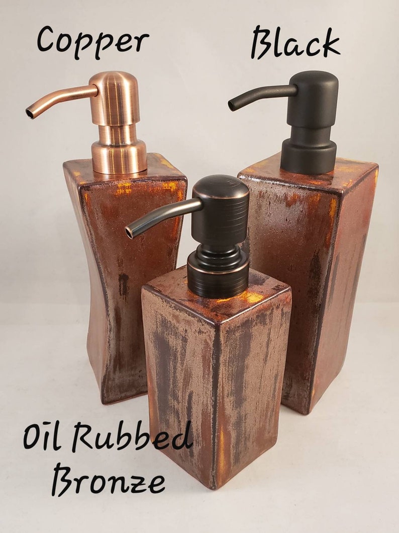 May include: Three decorative soap dispensers in copper, black, and oil-rubbed bronze finishes. The dispensers have square or rectangular bases and curved spouts. The word "Copper", "Black", and "Oil Rubbed Bronze" are written above the dispensers.