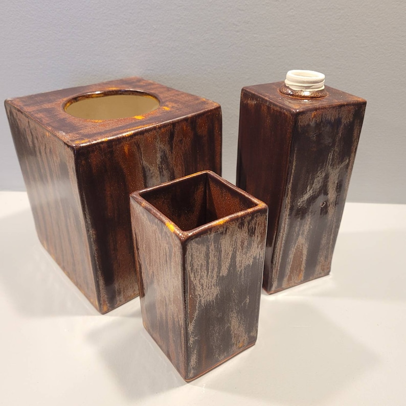 May include: A set of three ceramic bathroom accessories in shades of brown and bronze. The set includes a tissue box, a tall container with a lid, and a small square container. The pieces have a textured, wood-like appearance.