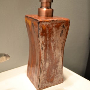 May include: A decorative soap dispenser with a copper-colored pump and a square, textured ceramic body. The ceramic has a brown and white mottled finish, adding a rustic touch to the bathroom accessory.