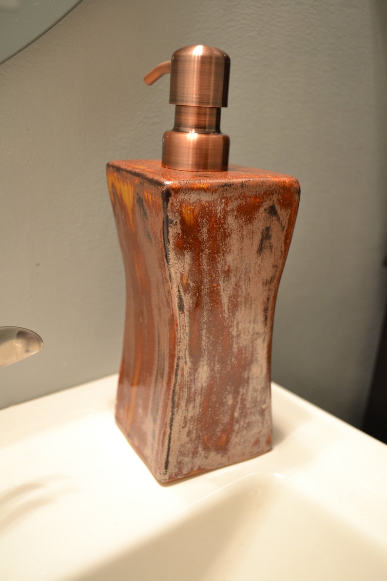 May include: A decorative soap dispenser with a copper-colored pump and a square, textured ceramic body. The ceramic has a brown and white mottled finish, adding a rustic touch to the bathroom accessory.