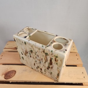 May include: A ceramic toothbrush holder with a speckled brown, green, and white glaze. It has four slots for toothbrushes and a rectangular compartment for toothpaste.