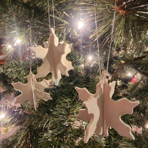 May include: Three white ceramic snowflake ornaments hanging on a green Christmas tree. The snowflakes are layered and have a subtle shimmer.