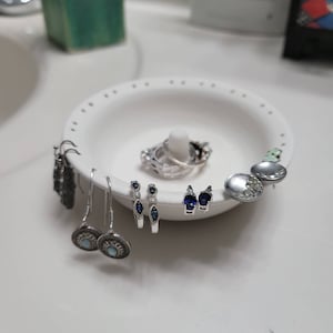 May include: A white ceramic dish with multiple holes, holding a variety of silver earrings with blue and clear gemstones. The earrings are arranged in a circle around a silver ring.