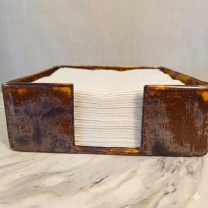 May include: A rectangular napkin holder with a distressed brown and gold finish. The holder is filled with a stack of white napkins. The design has a rustic, vintage aesthetic, suitable for kitchen or dining decor.