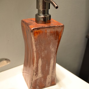 May include: A decorative soap dispenser with a bronze-colored pump and a textured, brown ceramic body. The dispenser has a rectangular shape with curved sides and a metallic finish, suitable for a bathroom.