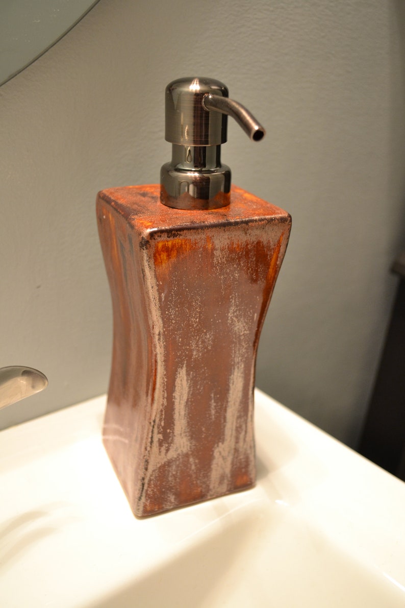 May include: A decorative soap dispenser with a bronze-colored pump and a textured, brown ceramic body. The dispenser has a rectangular shape with curved sides and a metallic finish, suitable for a bathroom.