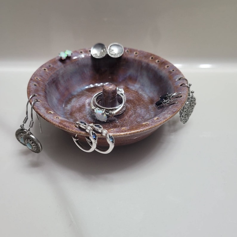 May include: A ceramic ring and earring holder with a purple and white glaze. The holder has multiple holes around the edge and a center post for rings. It is filled with various silver rings and earrings.