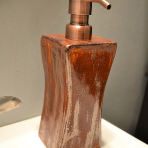 May include: A brown and copper-colored soap dispenser. The dispenser has a square, curved body with a copper-colored pump. The body has a textured, wood-like appearance. The dispenser is sitting on a white surface.