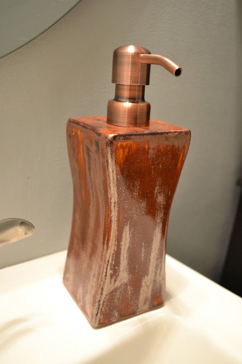 May include: A brown and copper-colored soap dispenser. The dispenser has a square, curved body with a copper-colored pump. The body has a textured, wood-like appearance. The dispenser is sitting on a white surface.