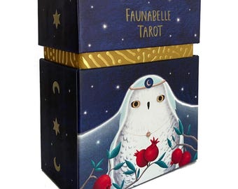 May include: A blue box with a gold trim and a white snowy owl with a crescent moon on its head. The box has the text "Faunabelle Tarot" on the top.