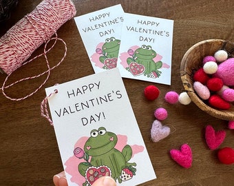 Peut inclure: Trois cartes imprimables pour la Saint-Valentin représentant une grenouille verte tenant une boîte de chocolats en forme de cœur. Les cartes ont un fond rose avec une bordure blanche et le texte "Joyeuse Saint-Valentin!" imprimé dessus.