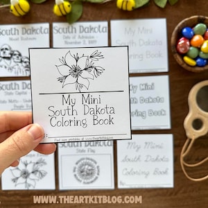 Puede incluir: Un libro para colorear en blanco y negro con el título "My Mini South Dakota Coloring Book". La portada presenta una ilustración en blanco y negro de una flor. El libro está sobre una mesa de madera con otras páginas para colorear y materiales de artesanía.