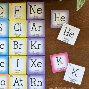 May include: Colorful square cards with the chemical symbols and names of elements from the periodic table. The cards are arranged in rows and columns, with each card featuring a different element. The cards are on a wooden surface.