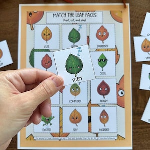 May include: A printable educational game titled "Match the Leaf Faces." The game features illustrated leaves with various expressions, including "Sleepy," "Cute," and "Surprised." A hand holds a leaf card with a sleepy face. The game is designed for print, cut, and play.
