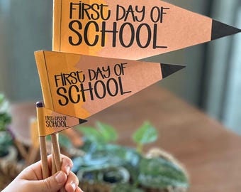 Op de afbeelding: Twee gele vlaggen in de vorm van een potlood met de tekst "First Day of School" op elk. De vlaggen zijn bevestigd aan houten potloden.