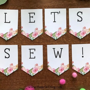 May include: A white banner with pink floral accents reads "Let's Sew!" in black lettering. The banner is made of paper and is perfect for a sewing party or craft event.