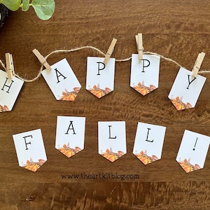May include: A white banner with the words "Happy Fall!" printed on it. The banner is strung with twine and clothespins, and each letter is on a separate piece of paper with a fall-themed design.