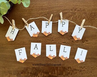 Könnte beinhalten: Ein weißes Banner mit den Worten "Happy Fall!" darauf gedruckt. Das Banner ist mit Schnur und Wäscheklammern befestigt, und jeder Buchstabe befindet sich auf einem separaten Blatt Papier mit einem Herbstmotiv.