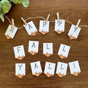 Puede incluir: Una guirnalda blanca con las palabras "HAPPY FALL Y'ALL!" en negro, cada letra en una banderola separada. Las banderolas están decoradas con gráficos temáticos de otoño y sujetas por pequeñas pinzas de madera en una cuerda. La guirnalda se muestra sobre una superficie de madera.