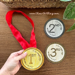 Puede incluir: Una mano sostiene una medalla dorada con una cinta roja, etiquetada como "1st". Se ven dos medallas adicionales, etiquetadas como "2nd" y "3rd". Las medallas tienen un diseño de corona de laurel y están sobre una superficie de madera.