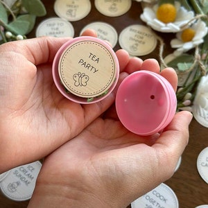 May include: A pink plastic container with a wooden lid. The lid has a butterfly design and the text "TEA PARTY".