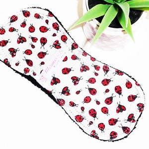 May include: A white reusable cloth menstrual pad with a black trim and a ladybug print. The pad has the text "Theme Sew" in pink lettering.