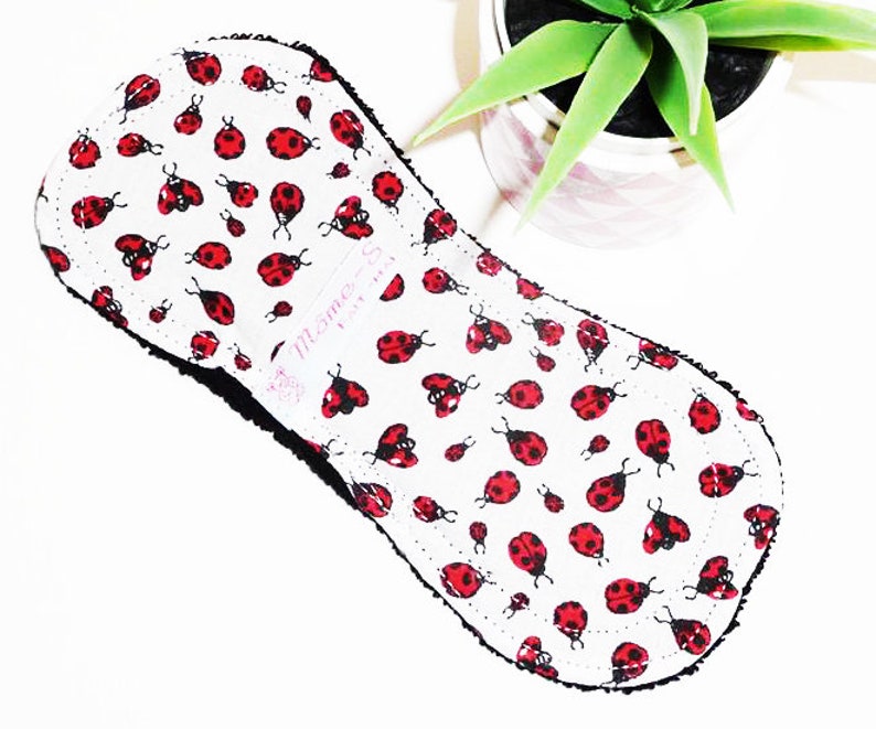May include: A white reusable cloth menstrual pad with a black trim and a ladybug print. The pad has the text "Theme Sew" in pink lettering.