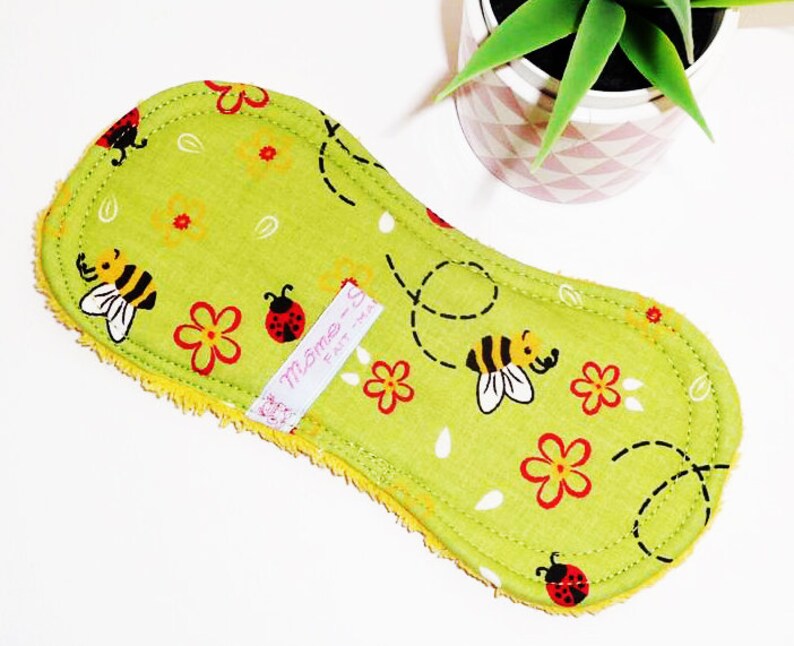 May include: Green reusable cloth pad with a bee and ladybug print. The pad has a yellow border and a white label that says "M&ocirc;me -  Fait &agrave; la main".