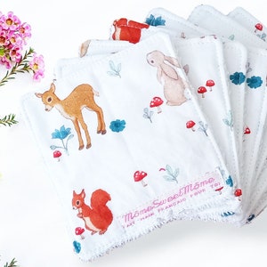 May include: A set of seven white cotton makeup remover pads with a woodland animal print. The print features a deer, a rabbit, a squirrel, mushrooms, and leaves. The pads have a white scalloped edge and the text "Mome Sweet Mome" in pink.